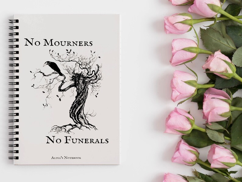 K&ouml;nnte beinhalten: Ein spiralgebundenes Notizbuch mit einer Schwarzwei&szlig;illustration eines Baumes mit einem Raben, der auf einem Ast sitzt. Der Text "No Mourners No Funerals Alina's Notebook" ist auf dem Cover gedruckt.