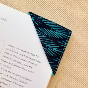 Peut inclure: Marque-page d'angle en tissu avec un motif bleu sarcelle et blanc sur fond bleu marine. Le marque-page est placé sur le coin d'un livre, avec les pages visibles. Le marque-page est en tissu et a des coutures blanches.