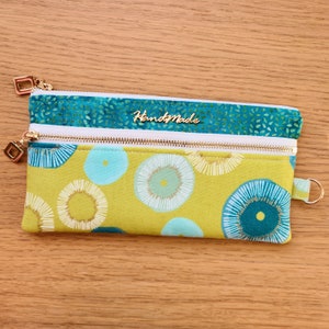 Può includere: Una pochette con cerniera in tessuto verde e blu con una cerniera bianca e accessori dorati. La pochette ha un motivo circolare e le parole "HandyMade" sono stampate sul davanti.