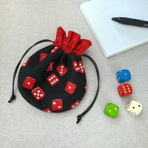 May include: A black fabric dice bag with red dice printed on it. The bag has a red drawstring and a black cord. There are five dice on a grey surface next to the bag.