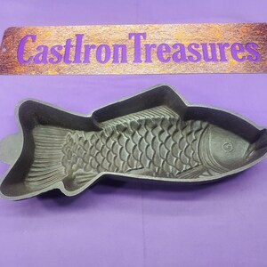 May include: A cast iron fish-shaped baking mold. The mold is gray and features detailed scales and fins. The text "CastIron Treasures" is above the mold on a wooden sign.