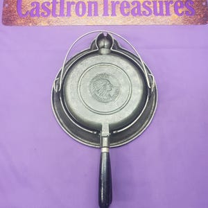 May include: A vintage cast iron waffle iron with a long, black wooden handle and a silver metal frame. The iron features a circular design with an embossed Native American profile and the text "Wapak Hollow Ware". The handle is attached to a hinge.
