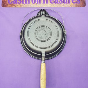 May include: A cast iron waffle iron with a wooden handle and a metal ring. The waffle iron has a round shape and a design of an American Indian head in the center.