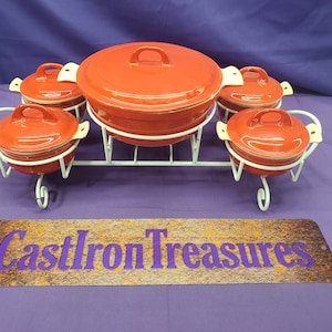May include: Set of five red enamel cast iron cookware with white lids on a white metal stand. The stand has a decorative scroll design. The set includes a large casserole dish and four smaller bowls. The set is labeled "Cast Iron Treasures".