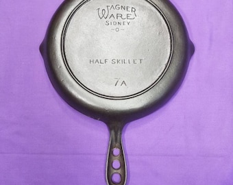 Puede incluir: Una sartén de hierro fundido negra a la mitad con un mango. La sartén está etiquetada "Wagner Ware Sidney -O- Half Skillet 7A".