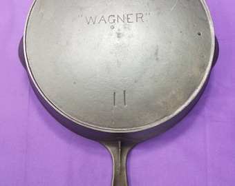 May include: A black cast iron skillet with a long handle. The skillet has the words "WAGNER" embossed on the bottom.