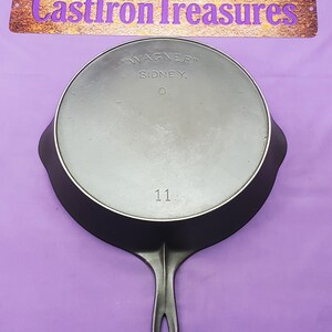 May include: A black cast iron skillet with a long handle, the bottom side facing up. The skillet is marked with "WAGNER" and "SIDNEY, O." and the number "11". The background is a solid purple color, with the text "CastIronTreasures" above the skillet.