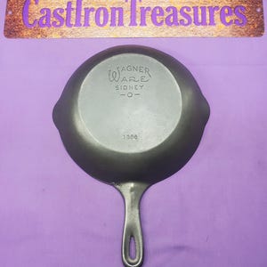 May include: A black cast iron skillet with the text "Wagner Ware Sidney -O- 1886" on the bottom. The skillet is on a purple surface.