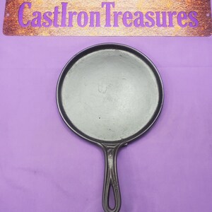 May include: A black cast iron griddle pan with a long handle and a circular cooking surface. The pan is centered on a purple background. Above the pan is a sign that reads "Cast Iron Treasures" in purple letters.
