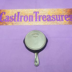 May include: A small, cast iron skillet with a handle, featuring the text "FAVORITE PIQUA" on the cooking surface. The skillet is set against a purple background, with a sign above that reads "CastIron Treasures".