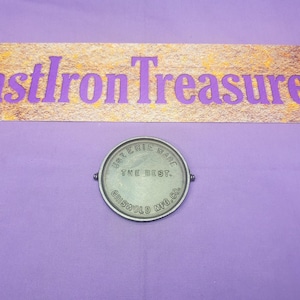 May include: A silver-colored cast iron cookware brand mark with the text "U.S. ERIE WARE THE BEST. GRISWOLD MFG. CO."