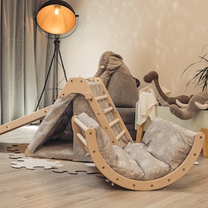 May include: A wooden play structure with a slide, climbing wall, and a rocking chair. The structure is painted in a natural wood color and has a gray fabric canopy. The play structure is in a room with a gray rug and a floor lamp.