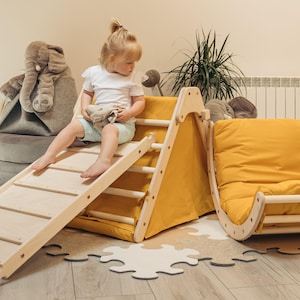 May include: A wooden climbing frame with a slide and a yellow fabric tent. A child is sitting on the slide. The frame is made of natural wood and has a yellow fabric covering. The slide is made of wood and has a yellow fabric covering. The tent is made of yellow fabric and has a wooden frame.