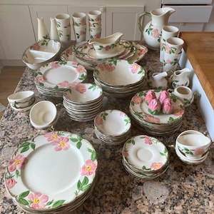 Peut inclure: Un ensemble de vaisselle en céramique blanche vintage avec un motif floral rose et des accents verts. L'ensemble comprend des assiettes, des bols, des tasses, des soucoupes, une saucière et un pichet. Les plats sont disposés sur un comptoir moucheté.