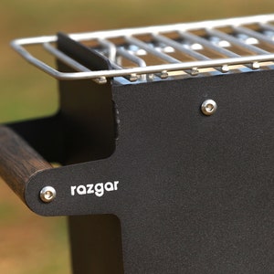 K&ouml;nnte beinhalten: Schwarzer Grill aus Metall mit einem Holzgriff und einem silbernen Grillrost. Der Grill hat den Schriftzug "razgar" auf der Seite.