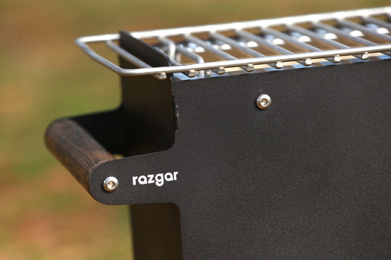 K&ouml;nnte beinhalten: Schwarzer Grill aus Metall mit einem Holzgriff und einem silbernen Grillrost. Der Grill hat den Schriftzug "razgar" auf der Seite.