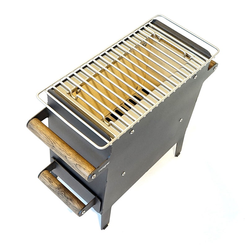 K&ouml;nnte beinhalten: Ein schwarzer Grill aus Metall mit einem Rost aus Edelstahl und Holzgriffen. Der Grill steht auf einem wei&szlig;en Hintergrund. Der Markenname "Ragnar" ist an der Seite sichtbar.