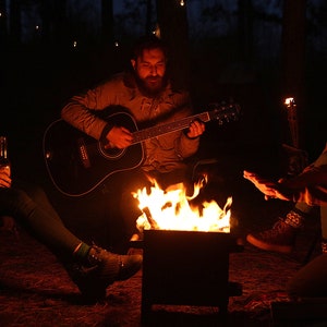 K&ouml;nnte beinhalten: Vier Personen sitzen um ein Lagerfeuer, einer von ihnen spielt eine Akustikgitarre. Das Feuer brennt hell in einer Metallfeuerstelle. Die Gruppe genie&szlig;t einen Abend in den W&auml;ldern.
