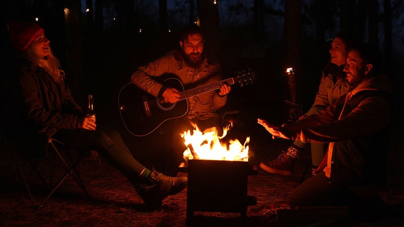 K&ouml;nnte beinhalten: Vier Personen sitzen um ein Lagerfeuer, einer von ihnen spielt eine Akustikgitarre. Das Feuer brennt hell in einer Metallfeuerstelle. Die Gruppe genie&szlig;t einen Abend in den W&auml;ldern.