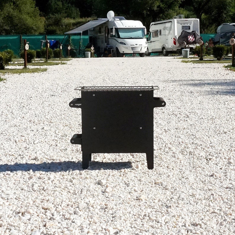 K&ouml;nnte beinhalten: Ein schwarzer, metallener Feuerplatz f&uuml;r den Au&szlig;enbereich mit einem Grillrost und dem Schriftzug "Ranger" an der Seite. Der Feuerplatz steht auf einer wei&szlig;en Kiesfl&auml;che.