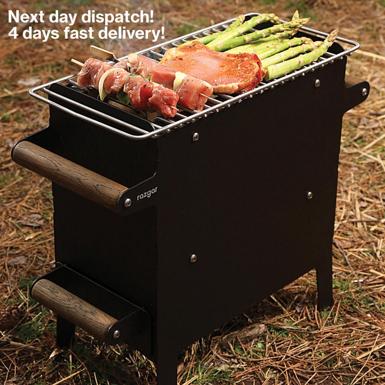 K&ouml;nnte beinhalten: Schwarzer Metall-Grill f&uuml;r den Au&szlig;enbereich mit Holzgriff und Edelstahlrost. Der Grill steht auf einem Bett aus Kiefernnadeln und es werden Spargel und Fleisch auf dem Rost gegrillt.