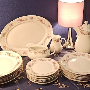 Może przedstawiać: Zestaw białej porcelanowej zastawy stołowej z kwiatowym wzorem. Zestaw zawiera dużą paterę, sosjerkę, dzbanek do śmietanki oraz kilka talerzy obiadowych i talerzy sałatkowych. Zastawa stołowa jest ułożona na niebieskim obrusie z złotym wzorem.