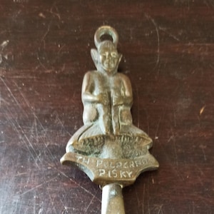 Könnte beinhalten: Ein Schuhlöffel aus Messing mit einer dekorativen Figur eines sitzenden Kobolds, der einen Schuh hält. Der Schuhlöffel trägt die Inschriften "In Polders" und "Pilsky".
