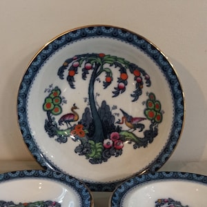 Może przedstawiać: Zestaw zabytkowych porcelanowych naczyń do serwowania. Zestaw zawiera dużą miskę i kilka mniejszych misek. Każdy element ma centralny wzór drzewa i ptaków z niebieską obwódką i złotym wykończeniem. Paleta kolorów obejmuje biel, błękit, zieleń, pomarańcz i złoto.