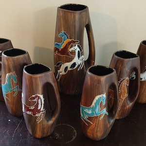 Puede incluir: Conjunto de siete tazas de cerámica marrón con un diseño de caballo estilizado en azul, blanco y rojo. Las tazas tienen un asa y están dispuestas en semicírculo sobre una superficie de madera.