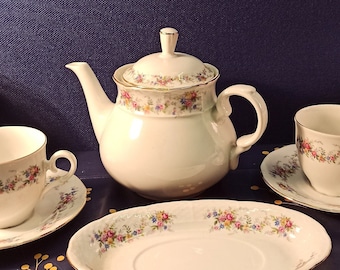 May include: A white porcelain tea set with floral patterns and gold trim. The set includes a teapot, two teacups with saucers, and a serving platter. The floral design features pink, blue, and yellow flowers. The set is arranged on a dark blue tablecloth.