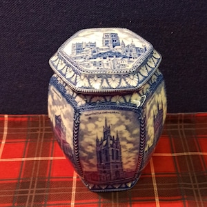 May include: A blue and white ceramic hexagonal jar with a lid. The jar is decorated with images of a cathedral and other buildings. The jar is sitting on a red and black tartan surface.