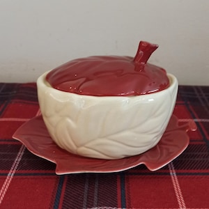 Peut inclure: Un bol à sucre en céramique en forme de pomme rouge et blanche avec un couvercle et une assiette en forme de feuille assortie. Le bol et l'assiette ont un fond blanc avec un motif de feuilles.