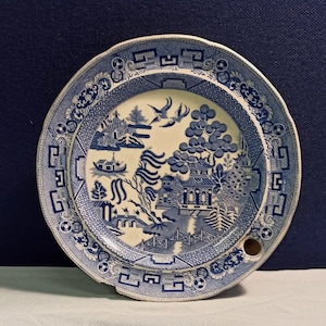 May include: A blue and white porcelain plate with a willow pattern design. The plate has a hole in the centre and a scalloped edge.