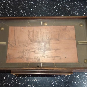 Puede incluir: Una caja rectangular de madera con una escena de paisaje grabada en color cobre bajo vidrio. La caja tiene herrajes de latón y un marco de madera oscura. La obra de arte representa una escena junto al agua con árboles y estructuras.