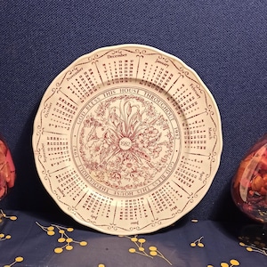 Könnte beinhalten: Ein weißer Keramikteller mit einem roten und braunen Blumendesign und einem Kalender für jeden Monat des Jahres. Der Teller trägt den Text "God Bless This House Throughout 1962" am Rand.