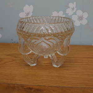 Może przedstawiać: Przezroczysta szklana miska z ozdobnym wzorem i trzema nóżkami. Miska stoi na drewnianej powierzchni.