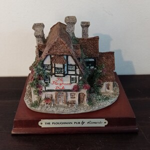 May include: A miniature replica of a pub called "The Ploughman Pub" by Leonardo. The pub is made of painted resin and features a brown roof, white walls, and a sign with the pub's name. The miniature is displayed on a wooden base.