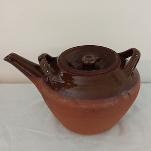 May include: A brown clay teapot with a lid and two handles. The teapot has a brown glaze on the top half and a natural clay finish on the bottom half.
