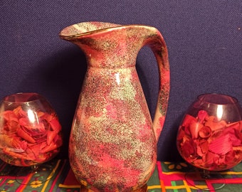 May include: A decorative ceramic pitcher with a speckled red and beige pattern, a gold base, and a curved handle. Two clear glass bowls filled with red potpourri sit on either side of the pitcher. The items are on a patterned tablecloth.