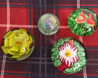 May include: Four glass paperweights with floral designs. One paperweight features a yellow flower, another has a red and white flower, and two have green leaves and flowers.