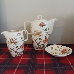 Könnte beinhalten: Eine weiße Teekanne, eine Milchkännchen und eine Zuckerdose aus Keramik mit einem Blumendesign. Die Teekanne hat einen Deckel und einen Ausguss. Die Milchkännchen und die Zuckerdose haben Griffe. Das Blumendesign zeigt rosa, gelbe und grüne Blumen auf weißem Hintergrund.