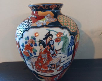 May include: A white ceramic vase with a blue, red and gold floral design. The vase features two women in traditional Japanese clothing sitting on a bench.