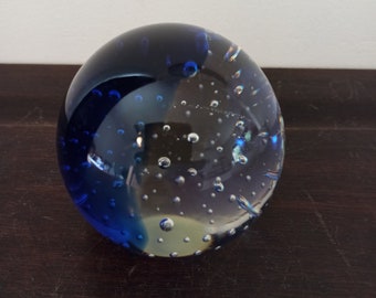 May include: A spherical glass paperweight. The top half is a deep blue, fading to clear with air bubbles. It rests on a dark brown surface. The paperweight is a decorative item.