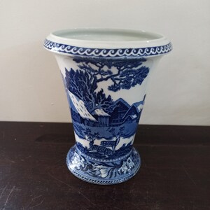 Puede incluir: Un jarrón de cerámica azul y blanco con un diseño floral y una escena de un pueblo de invierno con ciervos. El jarrón tiene un borde festoneado y una base acampanada.
