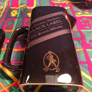 May include: Black ceramic Johnnie Walker Black Label Old Scotch Whisky decanter with a gold label and a walking man logo. The decanter has a handle and is sitting on a colorful patterned surface.