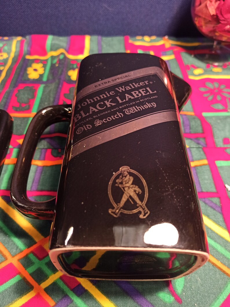 May include: Black ceramic Johnnie Walker Black Label Old Scotch Whisky decanter with a gold label and a walking man logo. The decanter has a handle and is sitting on a colorful patterned surface.