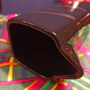May include: A black ceramic pitcher with a glossy finish. The pitcher has a wide opening and a rounded shape. The pitcher is sitting on a colorful fabric with a plaid pattern.