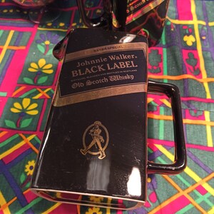 May include: Black ceramic decanter with a gold Johnnie Walker Black Label logo and a gold walking man logo. The decanter has a handle and is sitting on a colorful patterned fabric.