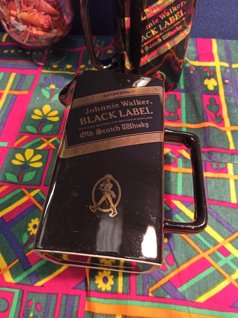 May include: Black ceramic decanter with a gold Johnnie Walker Black Label logo and a gold walking man logo. The decanter has a handle and is sitting on a colorful patterned fabric.