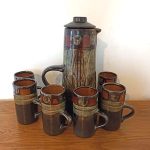 Può includere: Un set da caffè in ceramica con una caffettiera alta, marrone scuro con coperchio e sei tazze abbinate. Il set presenta un design strutturato e maculato con accenti marroni, arancioni e beige. Le tazze hanno manici.